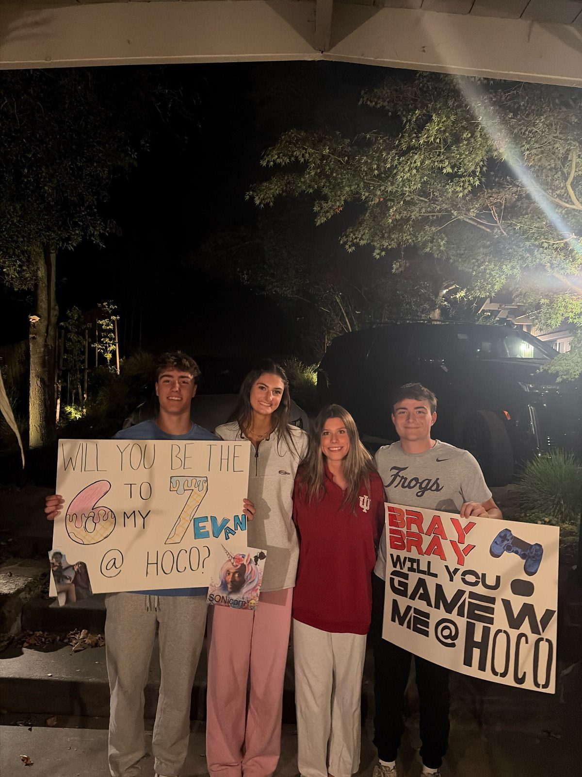 Four teenagers holding homecoming proposal posters while posing together outdoors at night.
