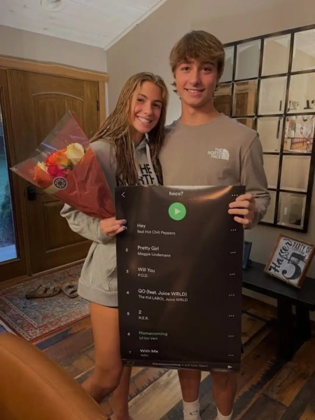 Teenage couple holding a playlist-themed homecoming proposal poster and a flower bouquet.
