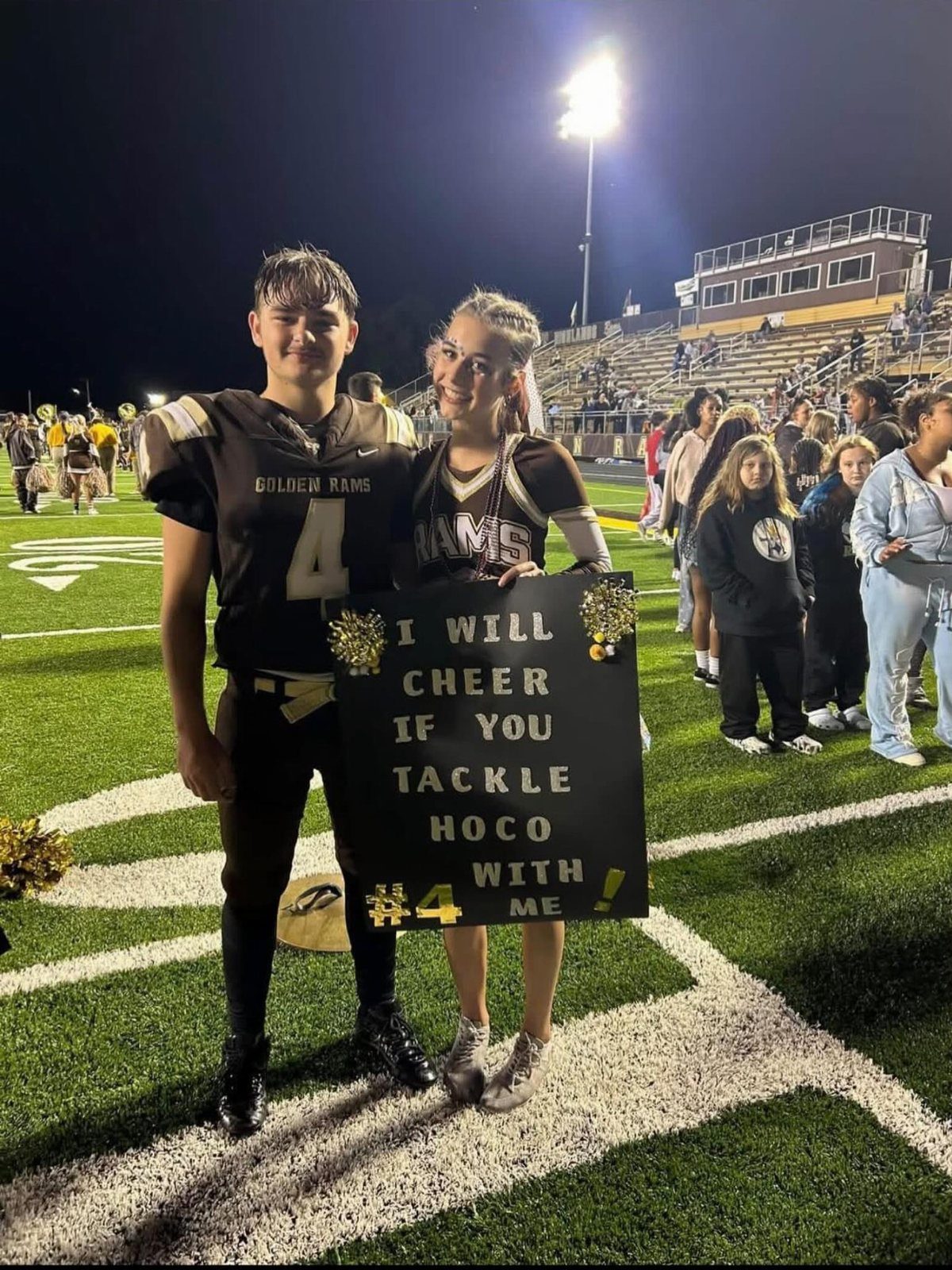 Football player and cheerleader posing on a field with a homecoming proposal sign.