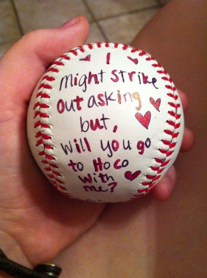 Baseball with handwritten homecoming proposal text and red hearts held in a hand.