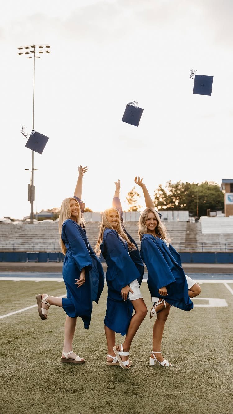 Three graduates in blue gowns tossing their caps in the air on a field