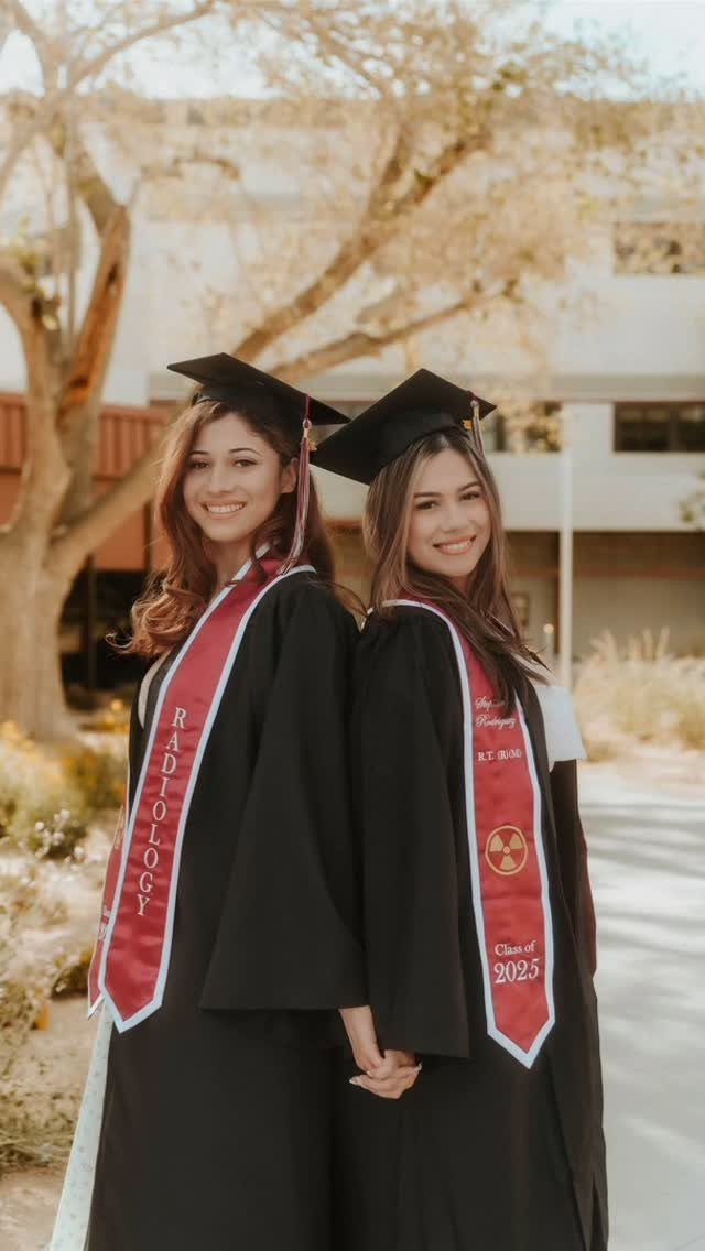 Two graduates in black caps and gowns with red sashes holding hands outdoors.