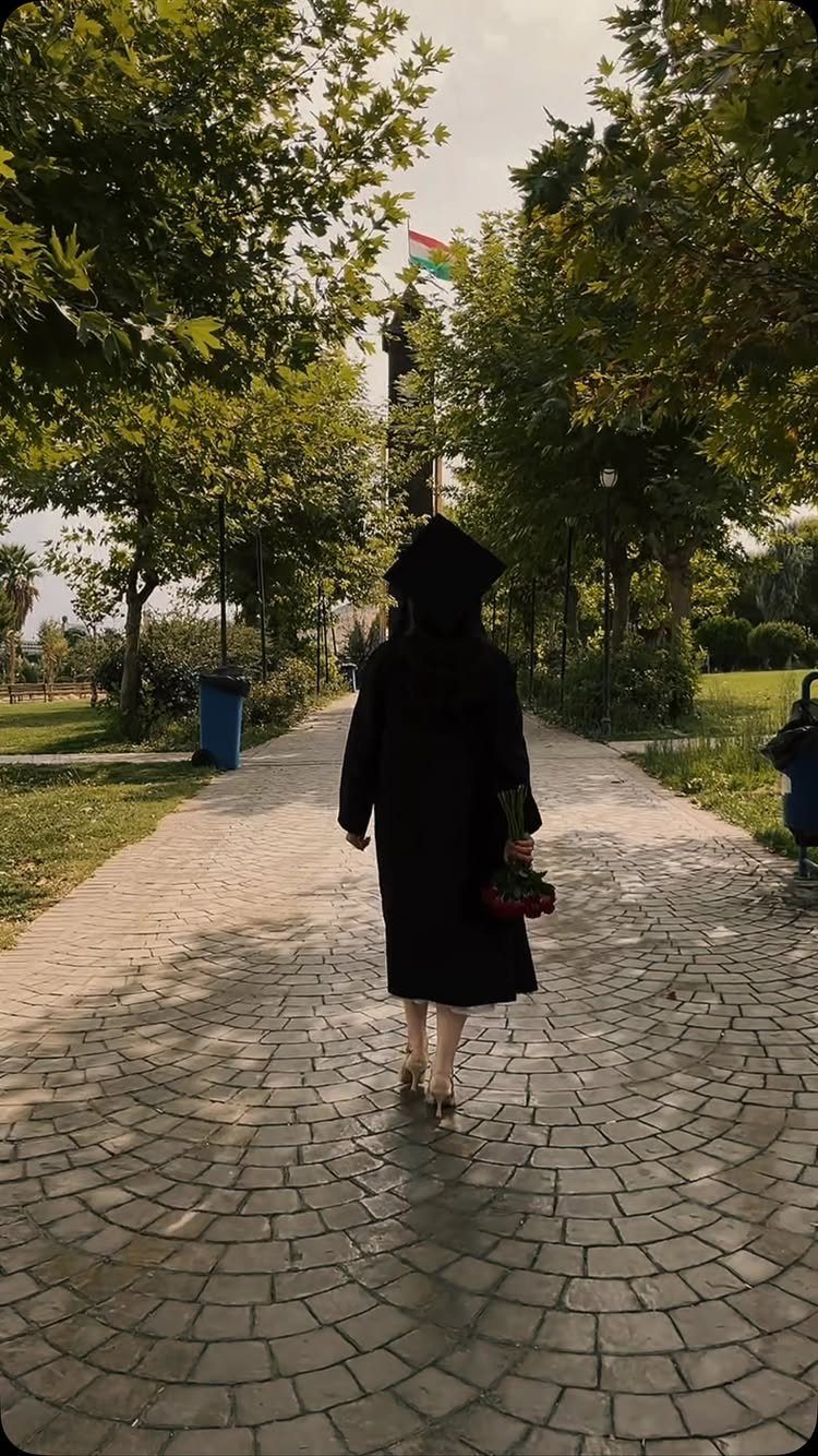 Graduate in black cap and gown walking away on a cobblestone path with roses.