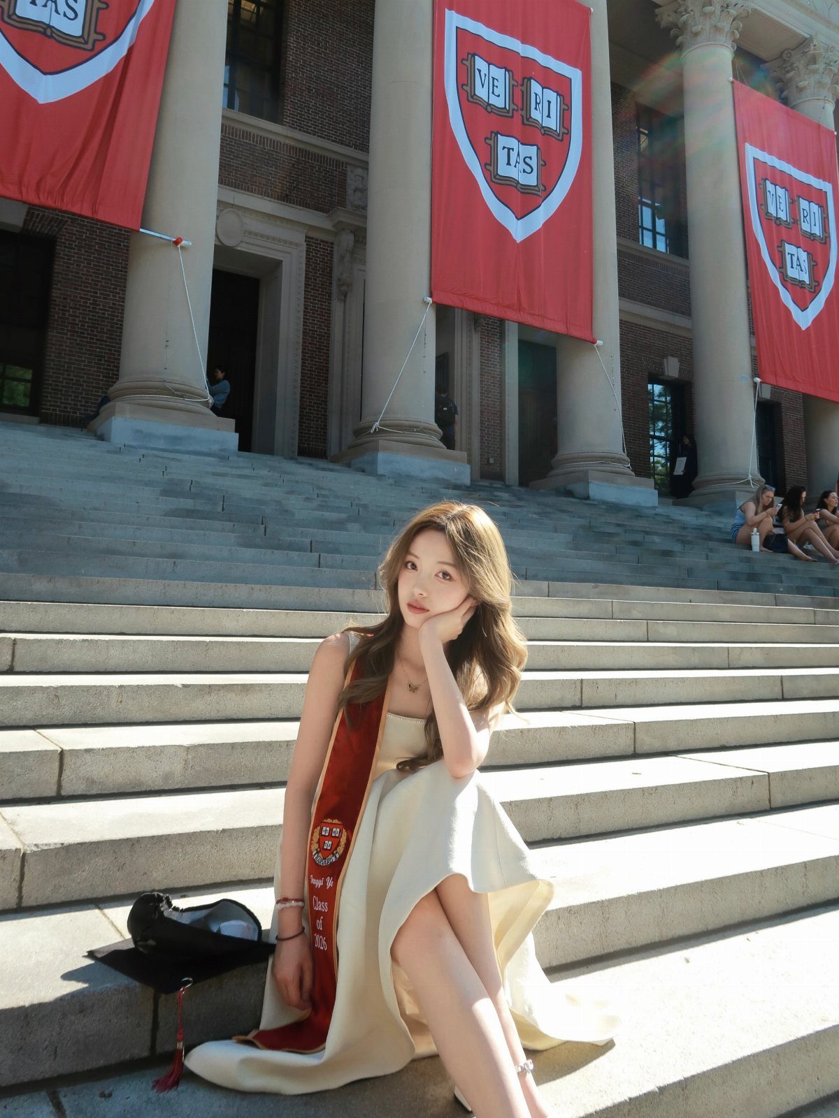 Young woman in a white dress and graduation stole sitting on stone steps.