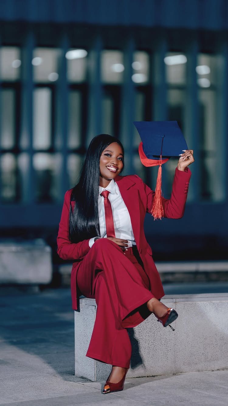 Woman in a red suit sitting on a ledge holding a graduation cap.