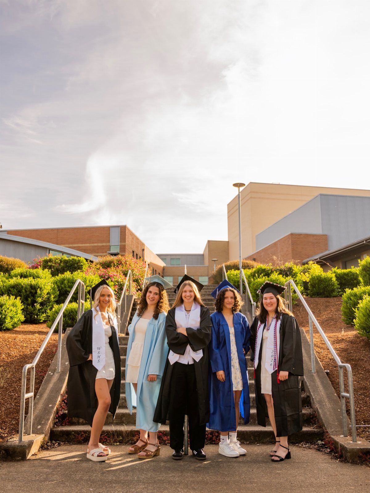 Five female graduates in caps and gowns posing together on outdoor school stairs.