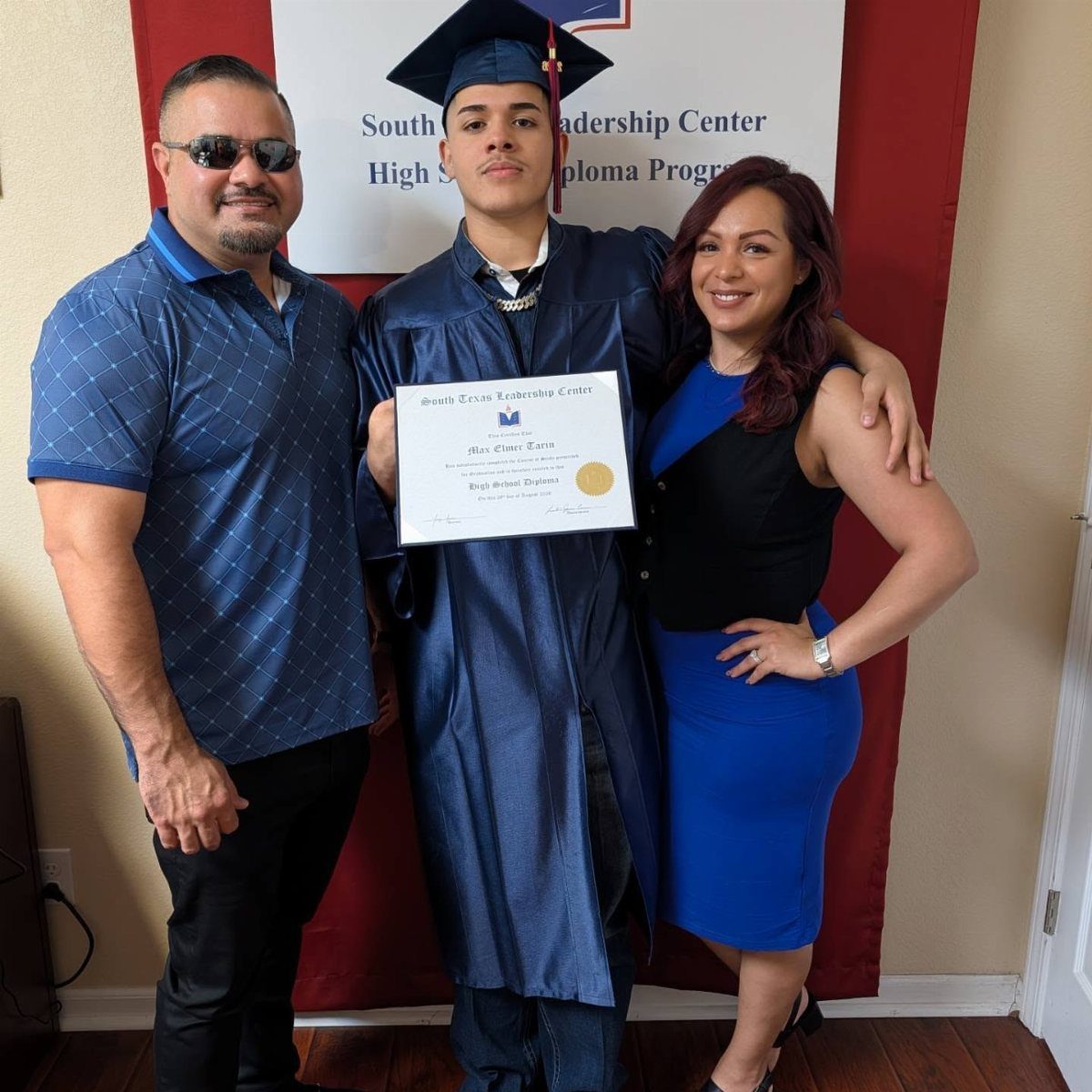 Graduate in blue cap and gown posing with parents holding a diploma certificate.