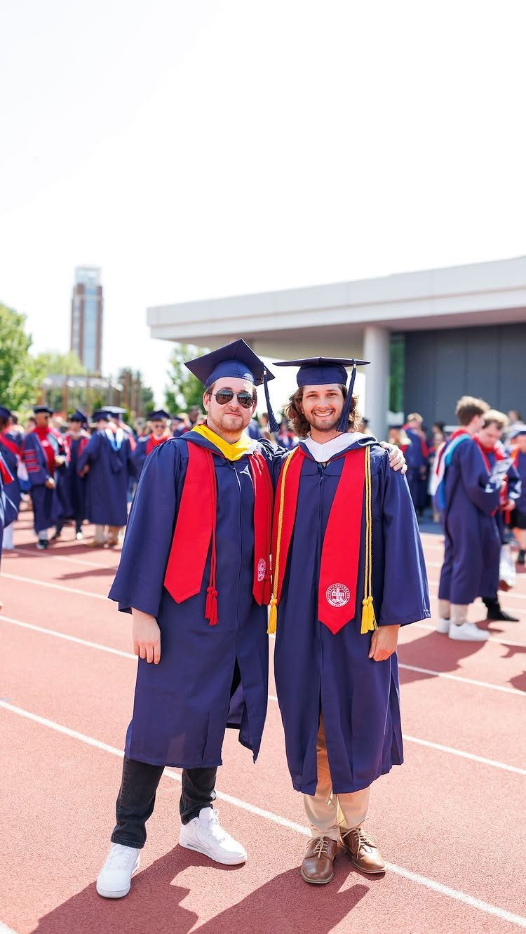 Two graduates in navy gowns and red stoles posing on a red running track.