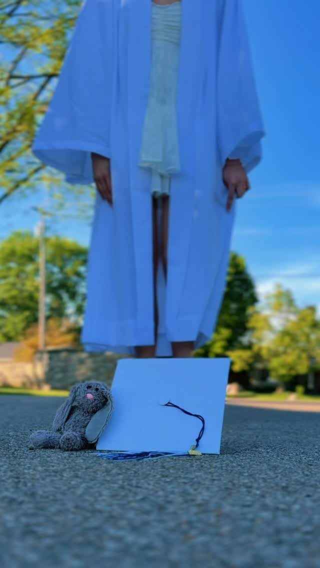 White graduation cap and grey plush bunny on asphalt with blurred graduate.