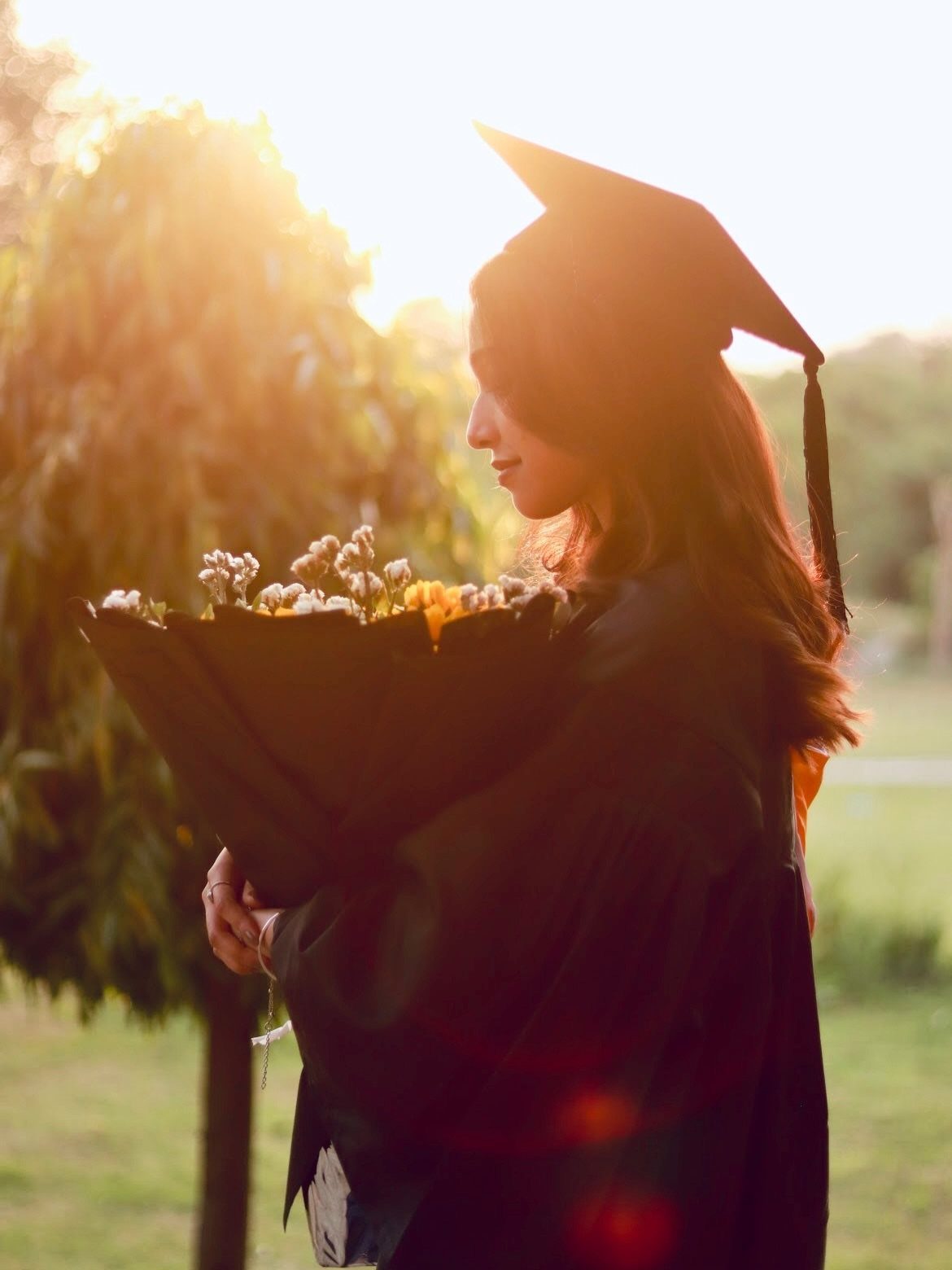 Graduate in cap and gown holding flowers during a golden hour sunset.