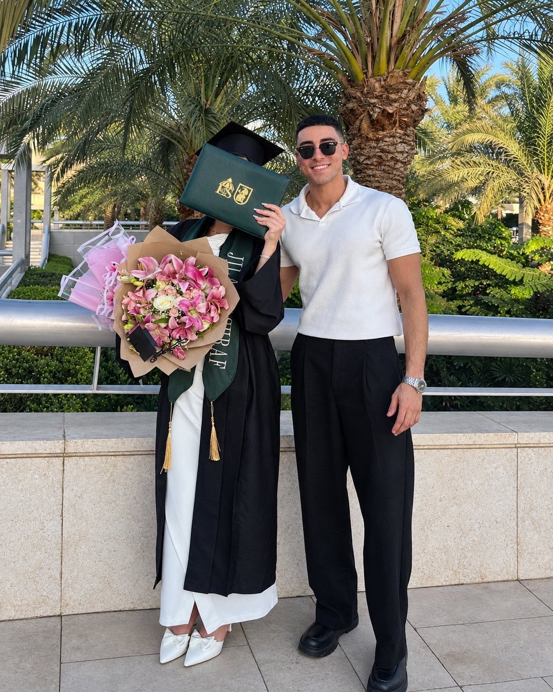 Graduating woman in black gown and man in white polo posing together outdoors.