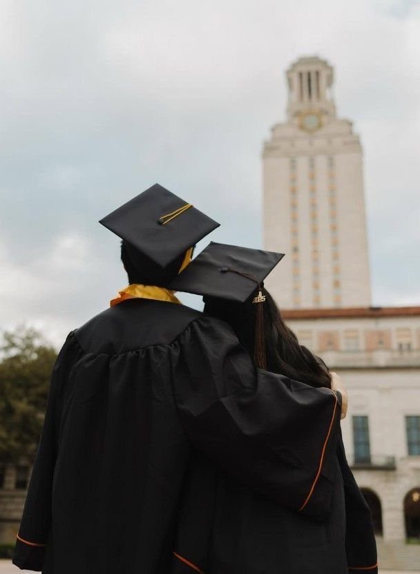 Couple in black graduation gowns and caps embracing before a university tower