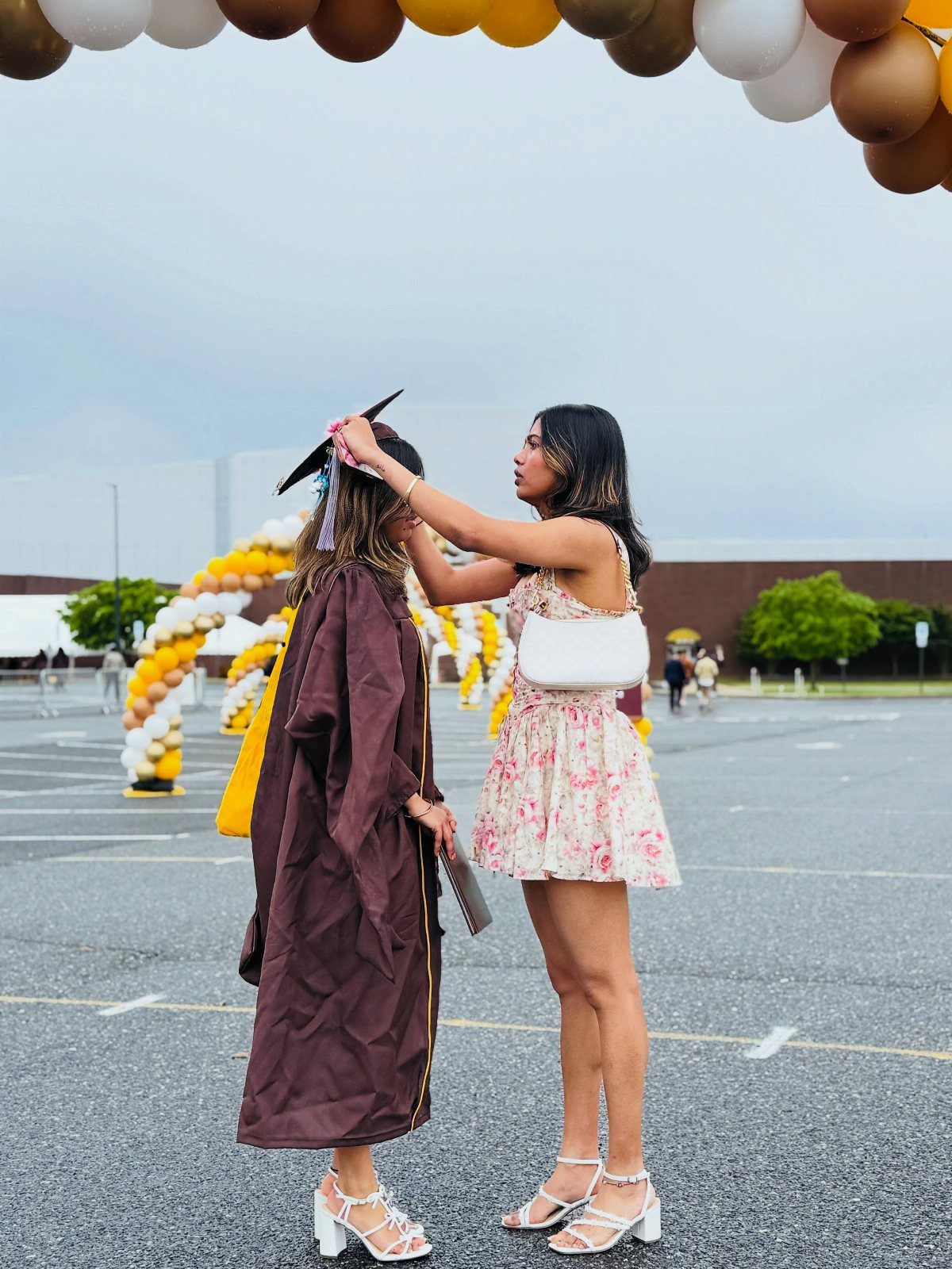 Woman in floral dress adjusting graduation cap on woman in brown gown