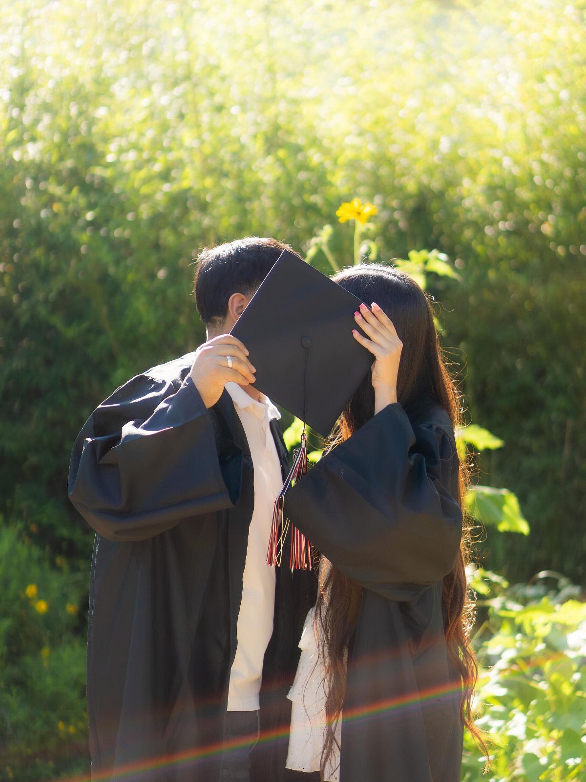 Couple in black graduation gowns holding a cap to hide their faces outdoors.