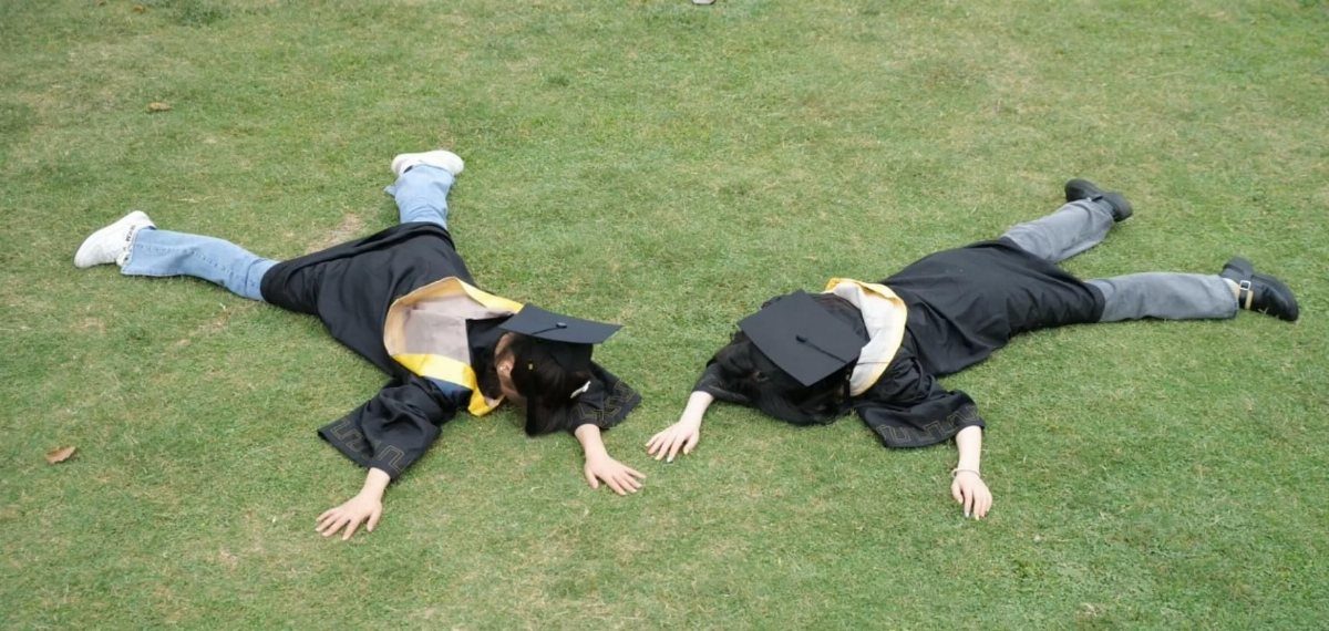 Two graduates in black gowns and caps lying on a green grass lawn.