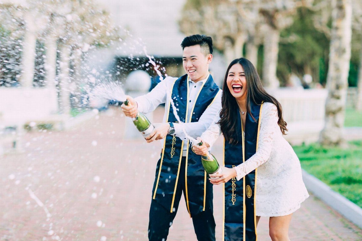 Graduating couple laughing while popping champagne bottles outdoors on a campus path.