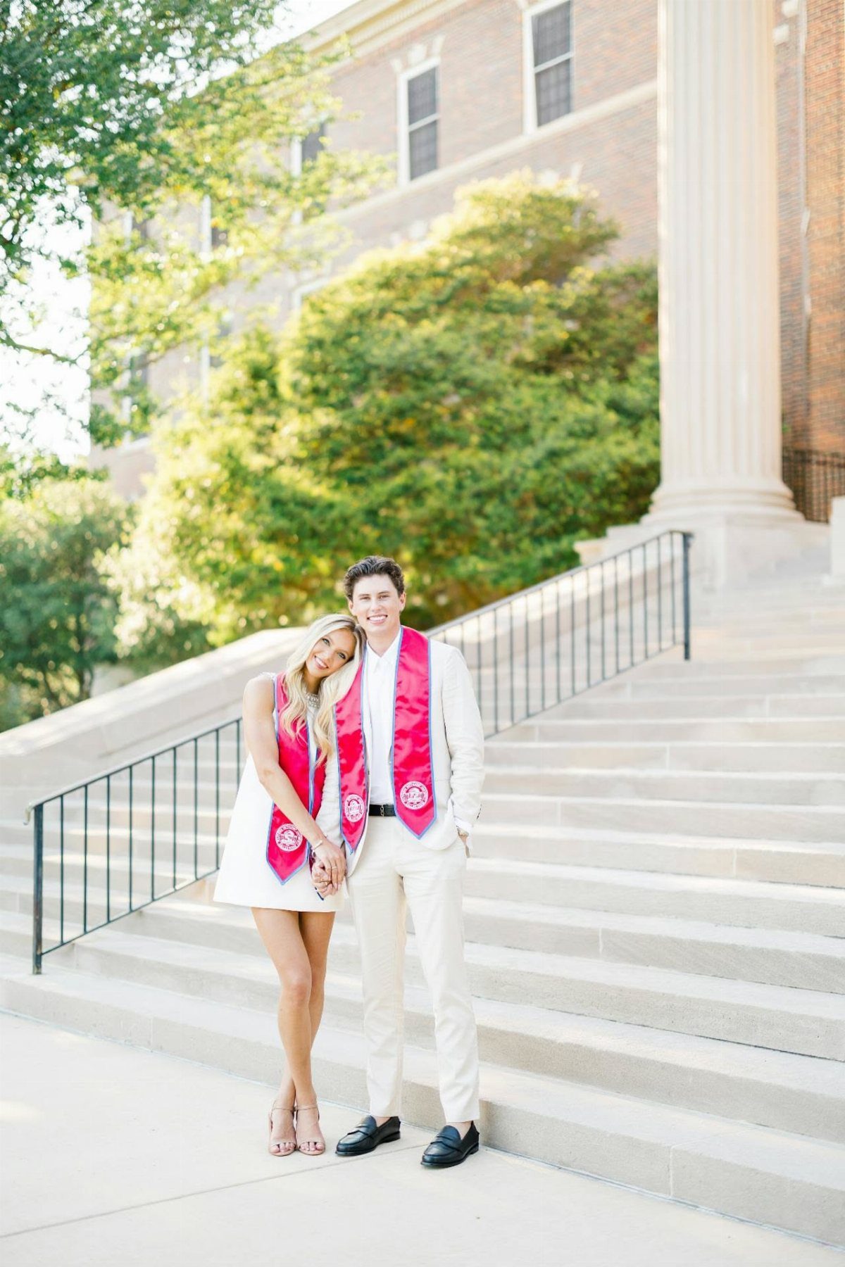 Couple in white outfits and red graduation stoles posing on campus stairs.