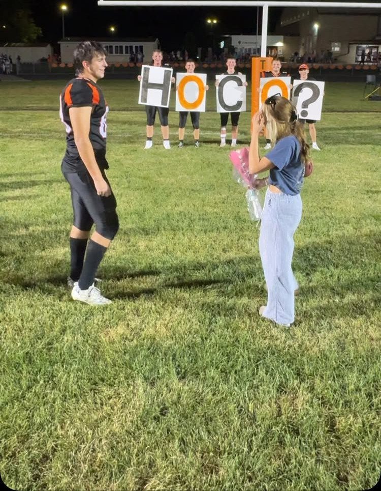 Football player proposing to a girl with friends holding HOCO signs on a field