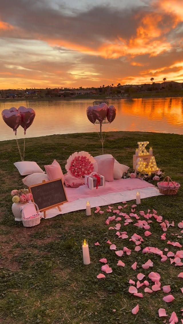 Romantic picnic setup with pink petals, heart balloons, and candles by a lake.