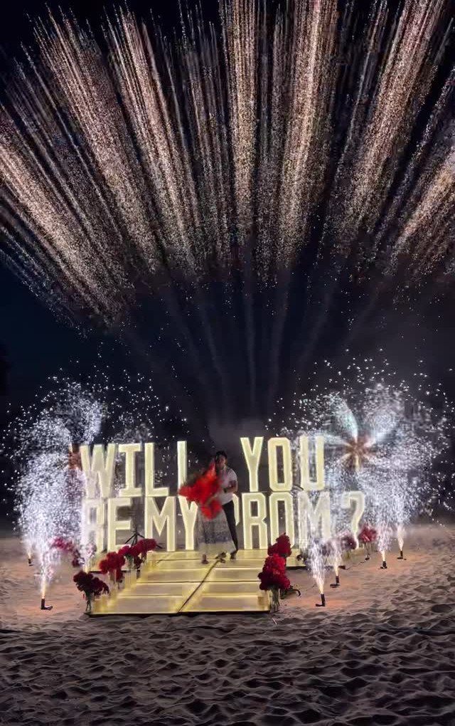 Couple standing before large glowing letters asking for a prom date with fireworks.