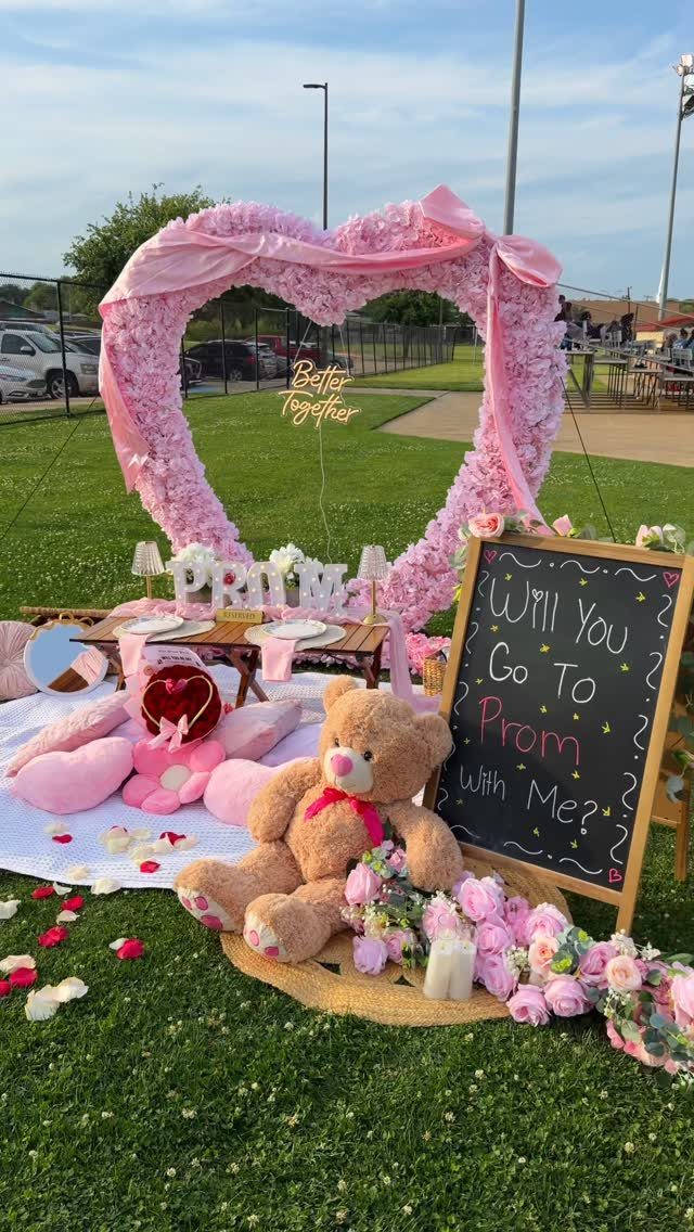 Promposal setup with a large pink floral heart, teddy bear, and chalkboard sign.