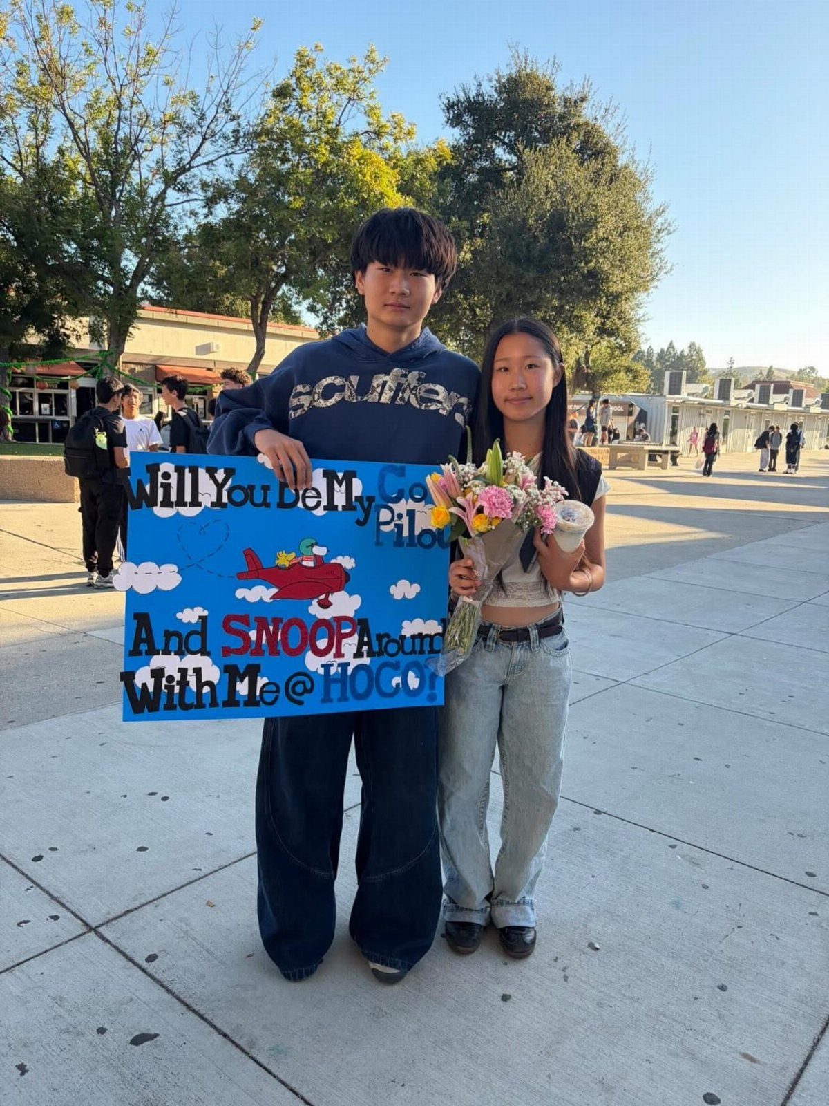 Teenage couple posing with a blue Snoopy themed promposal poster and flowers outdoors.