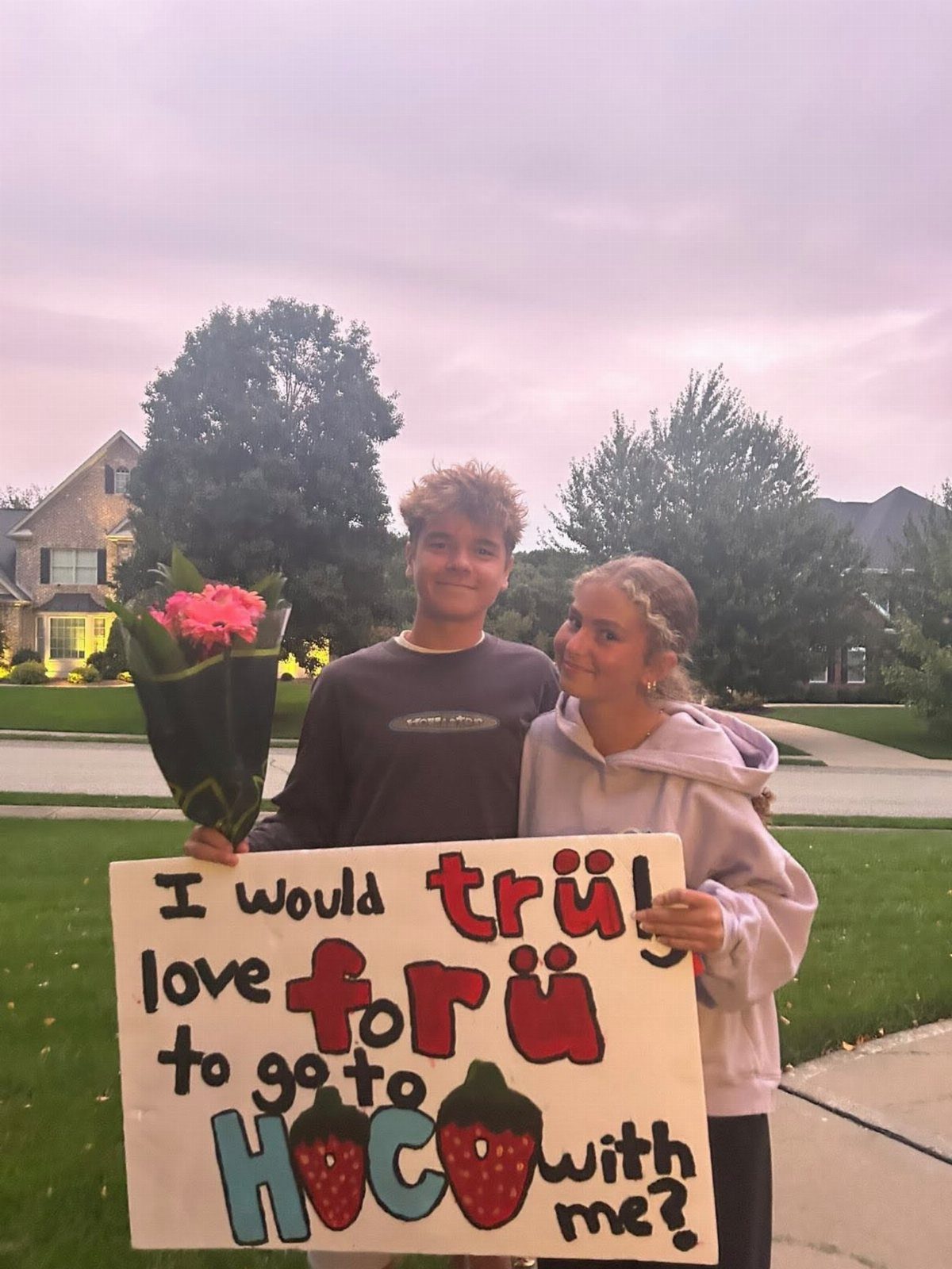 Teenage couple holding a berry themed homecoming proposal poster and pink flowers outdoors.