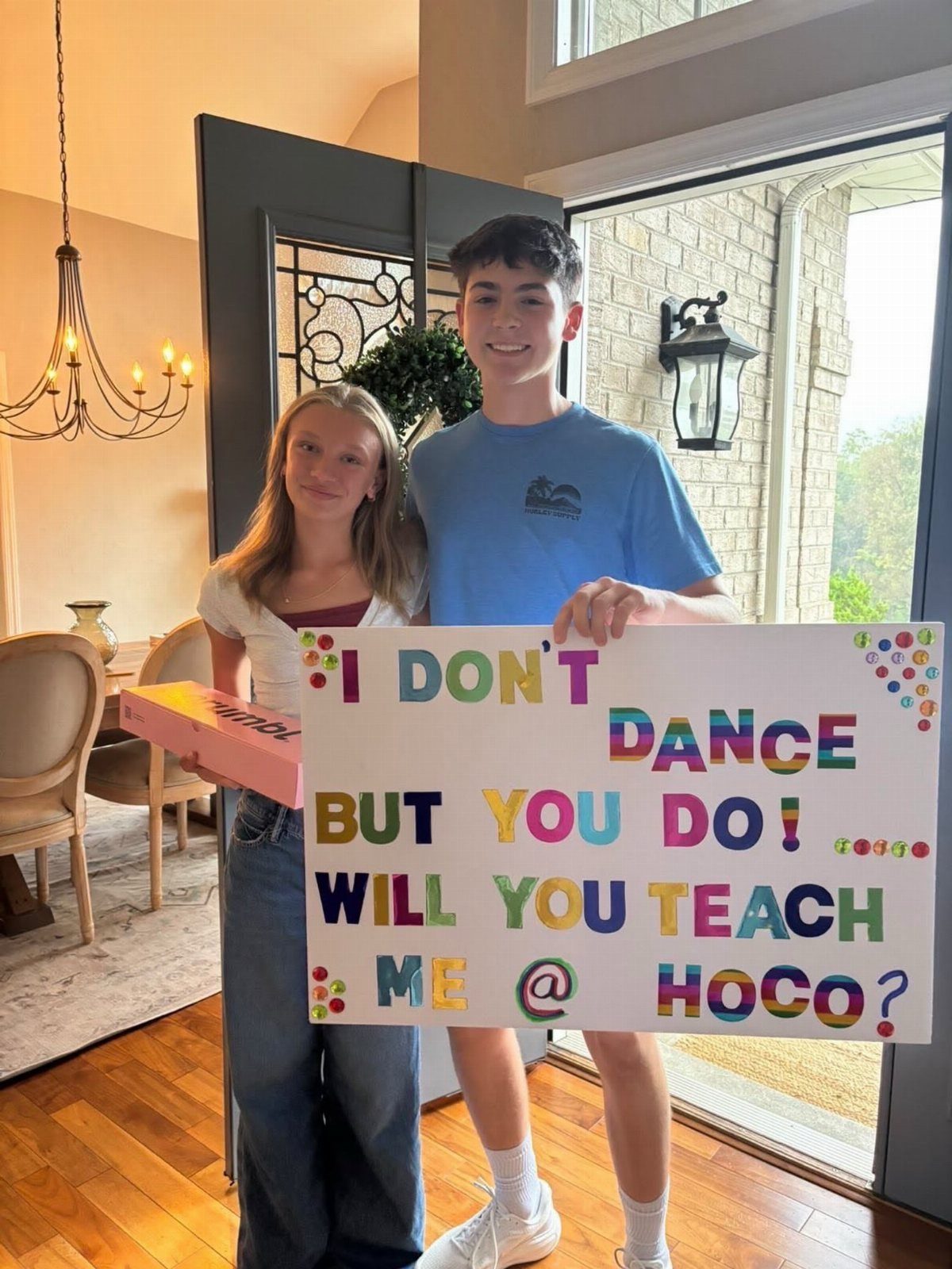 Teenage boy holding a dance themed homecoming proposal poster next to a girl.