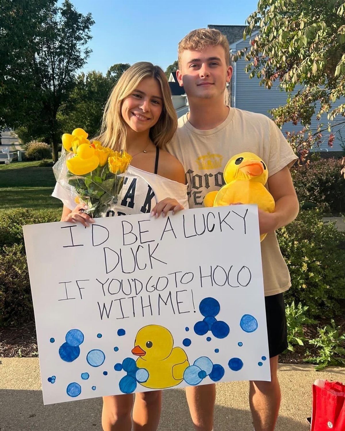 Teenage couple posing with a duck-themed homecoming proposal poster and yellow roses.