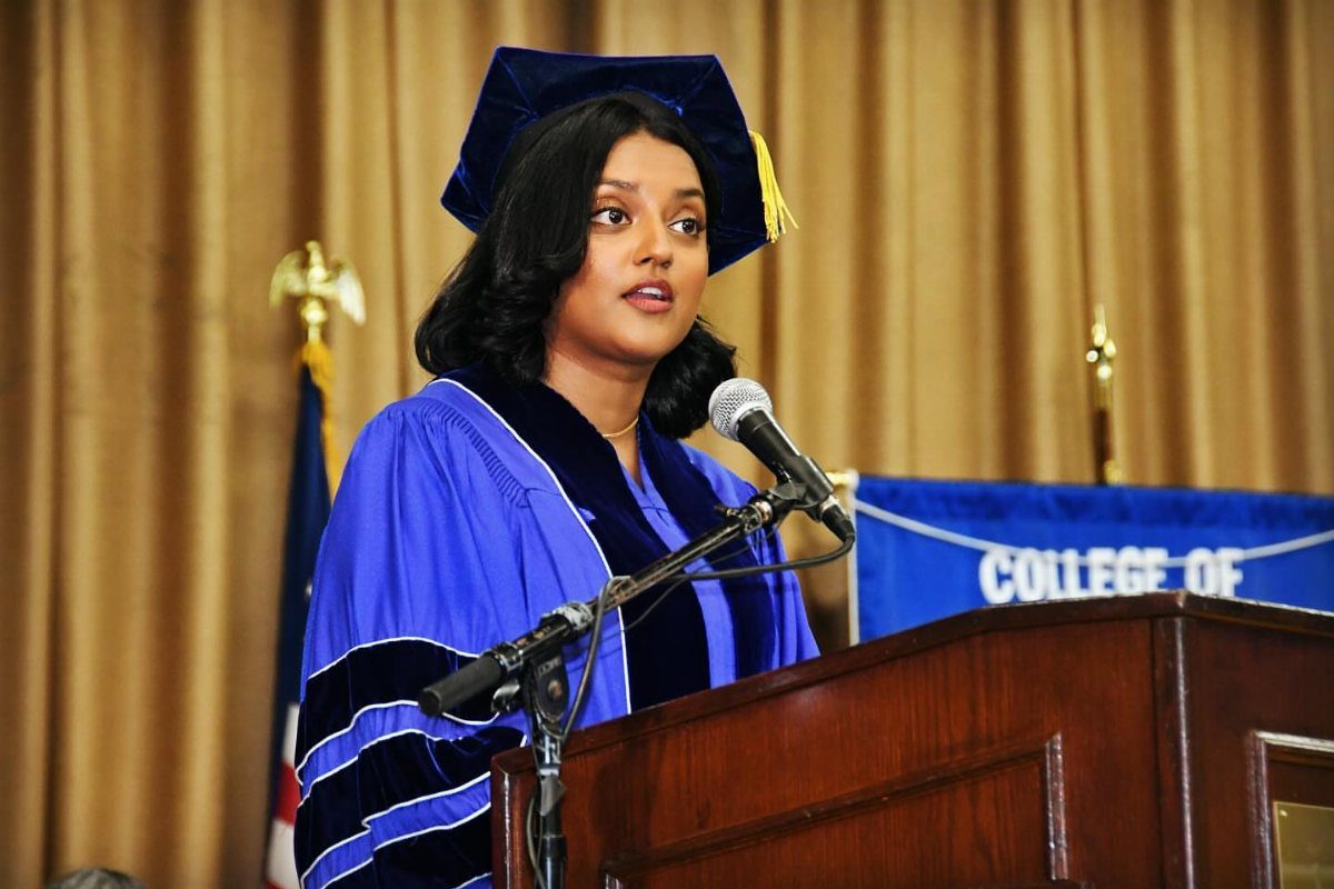 Woman in blue doctoral regalia speaking at a podium during a graduation ceremony.