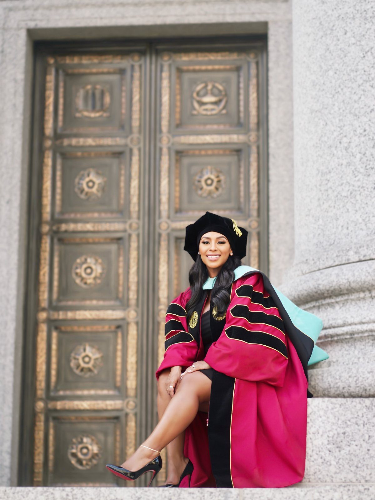 Woman in red doctoral regalia and light blue hood sitting by bronze doors.