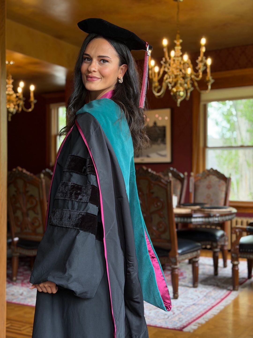 Woman wearing a black doctoral tam, gown, and teal and pink academic hood.