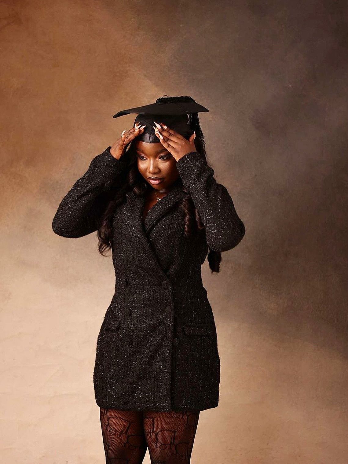 Woman in a black blazer dress and graduation cap posing against brown background