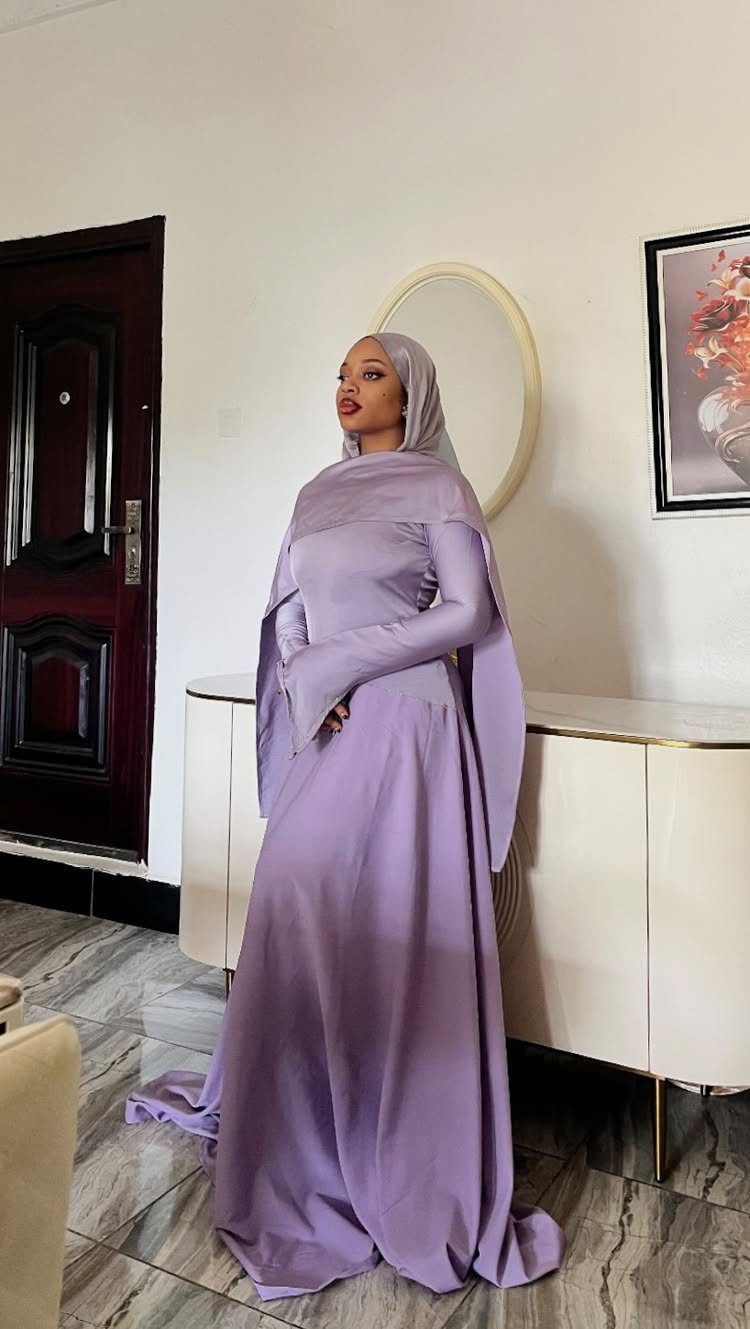 Woman in a long-sleeved lavender purple gown and matching hijab standing indoors.
