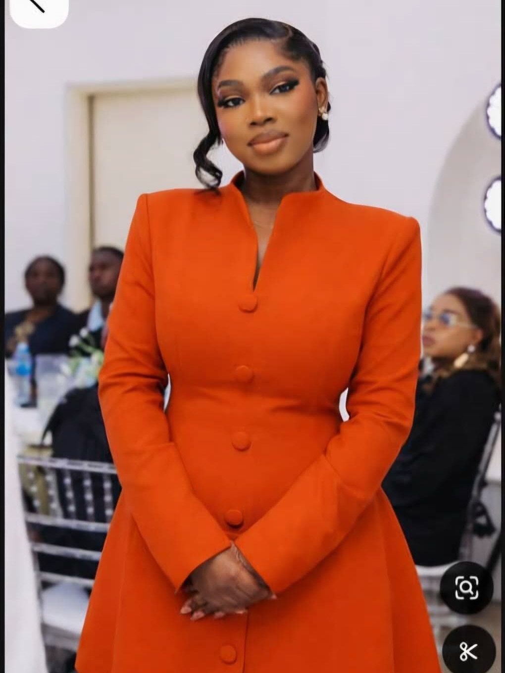 Woman wearing a structured orange long-sleeved dress with buttons and a high collar.