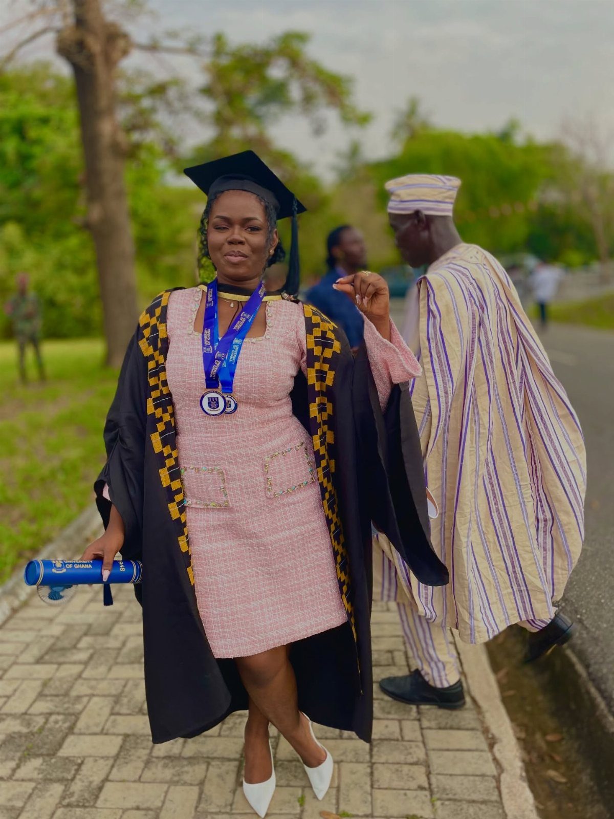Woman in a pink dress and graduation gown holding a blue diploma outdoors.