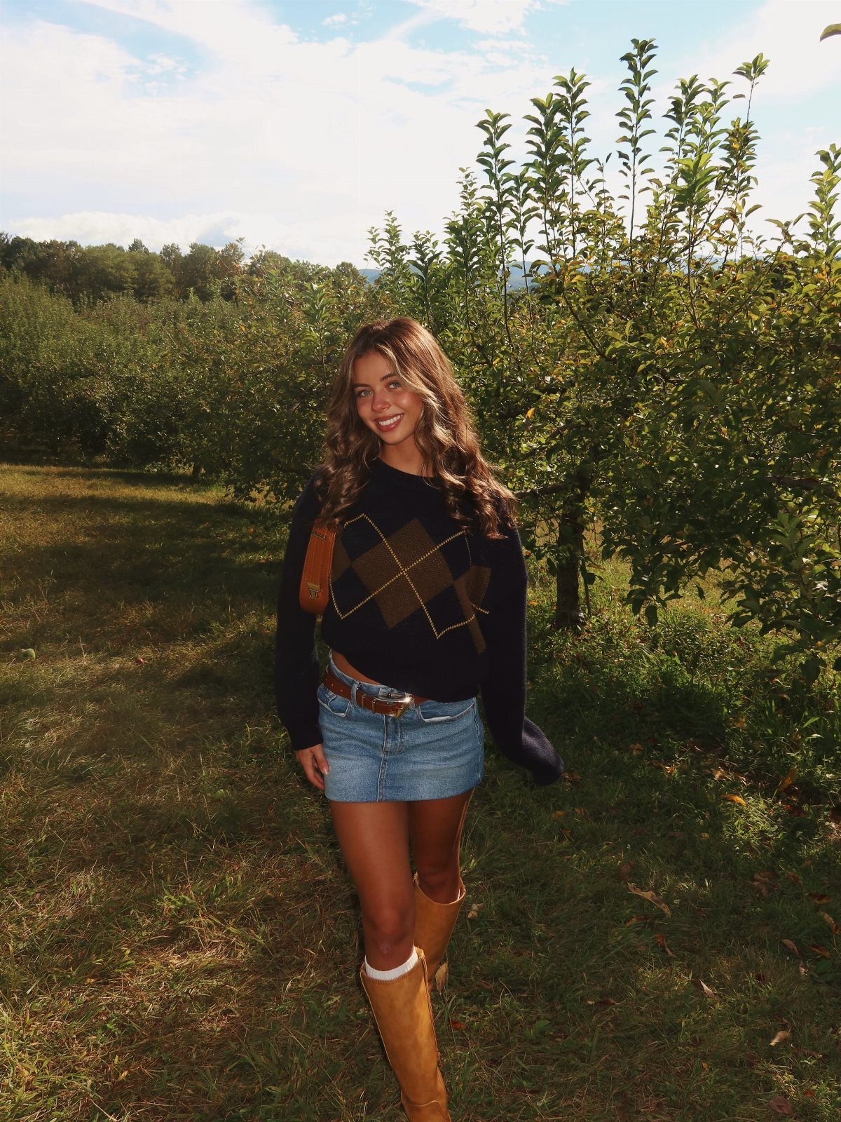 Young woman in argyle sweater and denim skirt posing in a sunny apple orchard