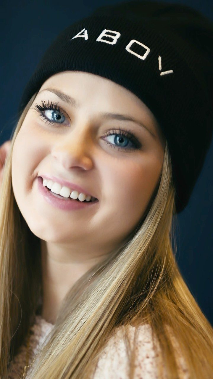 Smiling young woman wearing a black knit beanie with white text.