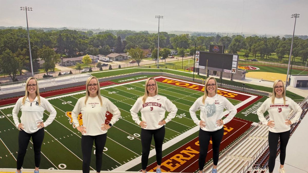 Five women in white tops and black leggings posing at a football stadium