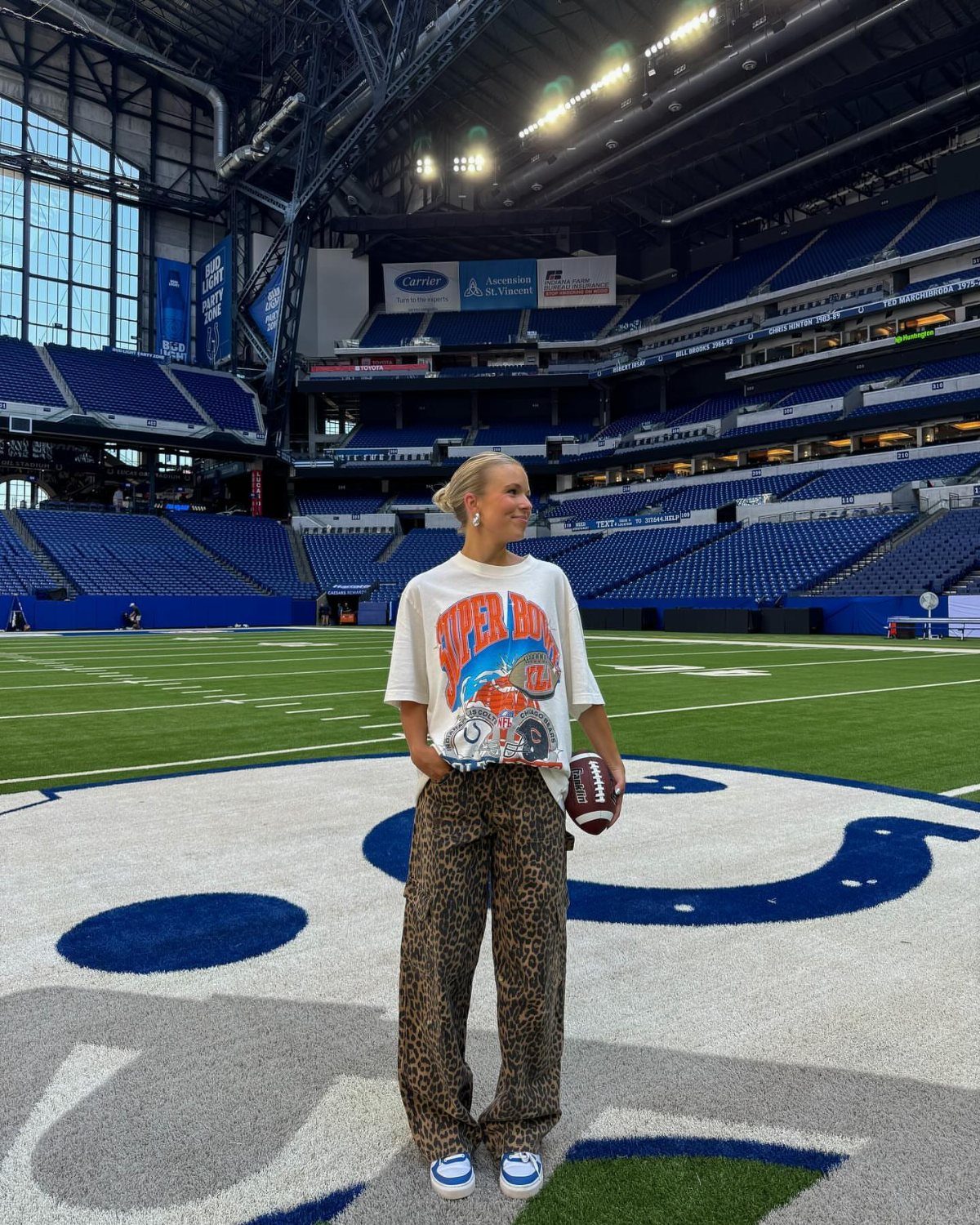 Woman in white graphic tee and leopard pants holding a football in a stadium