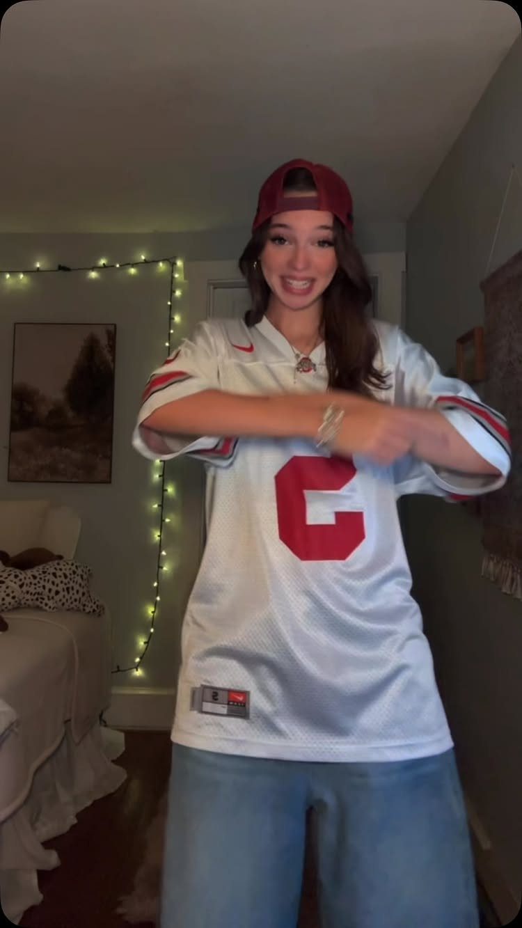 Woman wearing an oversized white football jersey and light blue jeans in a bedroom.