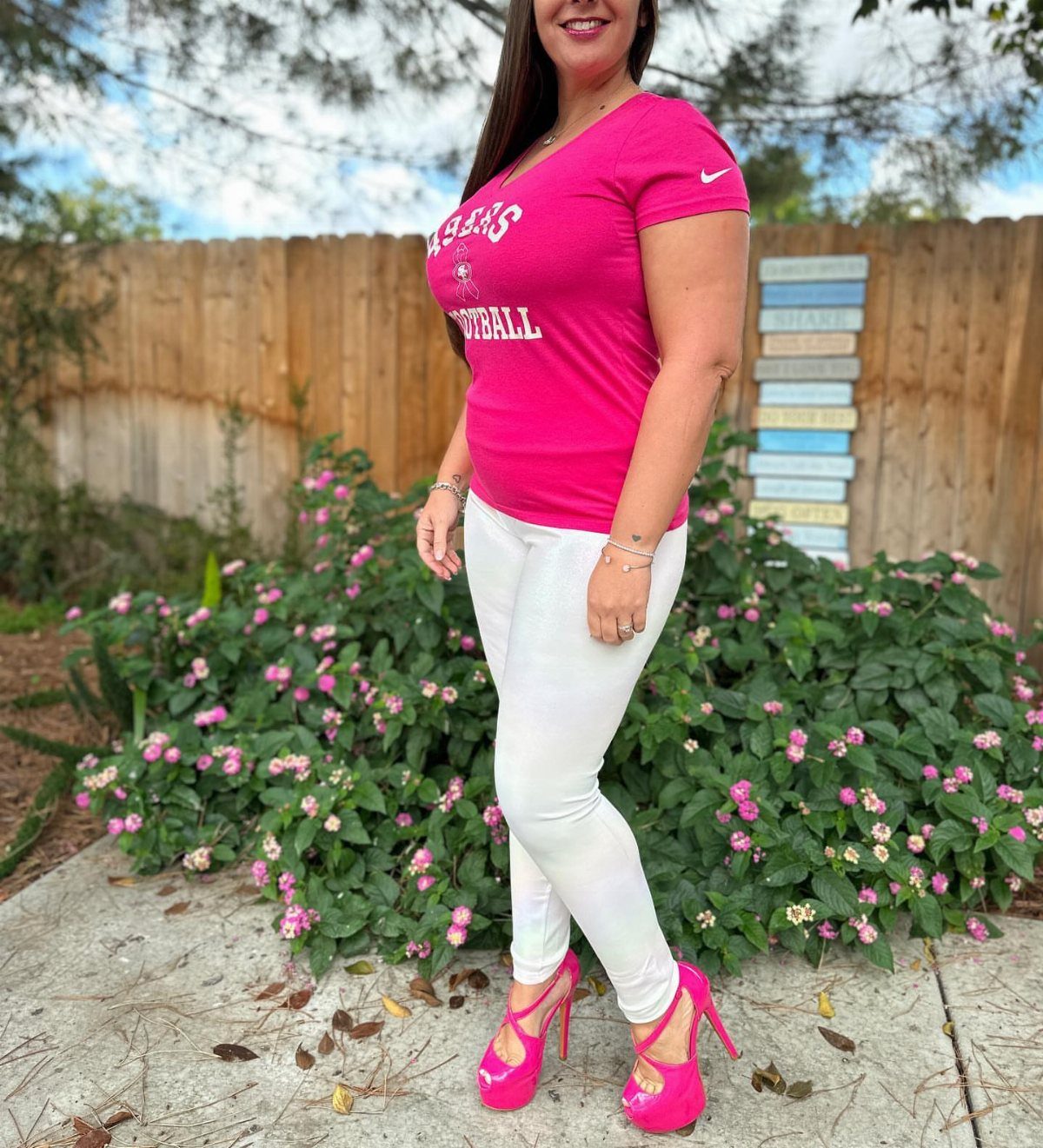 Woman in pink football t-shirt, white pants, and pink high heels outdoors.