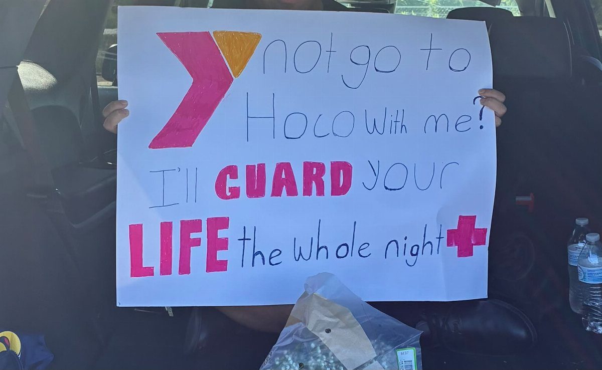 Handmade homecoming proposal poster held by a person inside a dark car interior.
