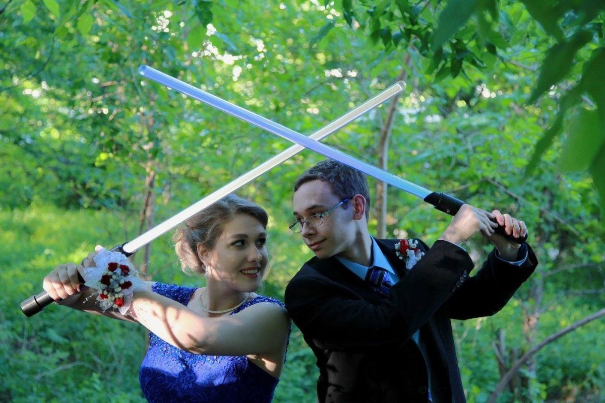 Couple in formal wear crossing glowing white toy swords in a green forest