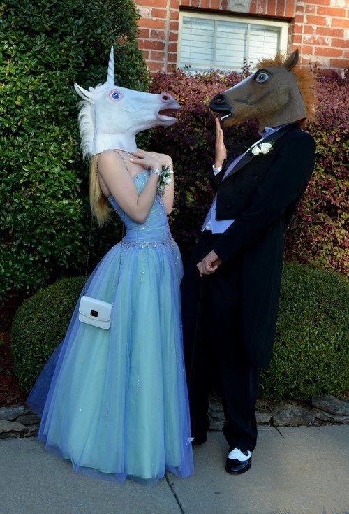 Couple in prom attire wearing unicorn and horse masks posing outdoors near bushes.