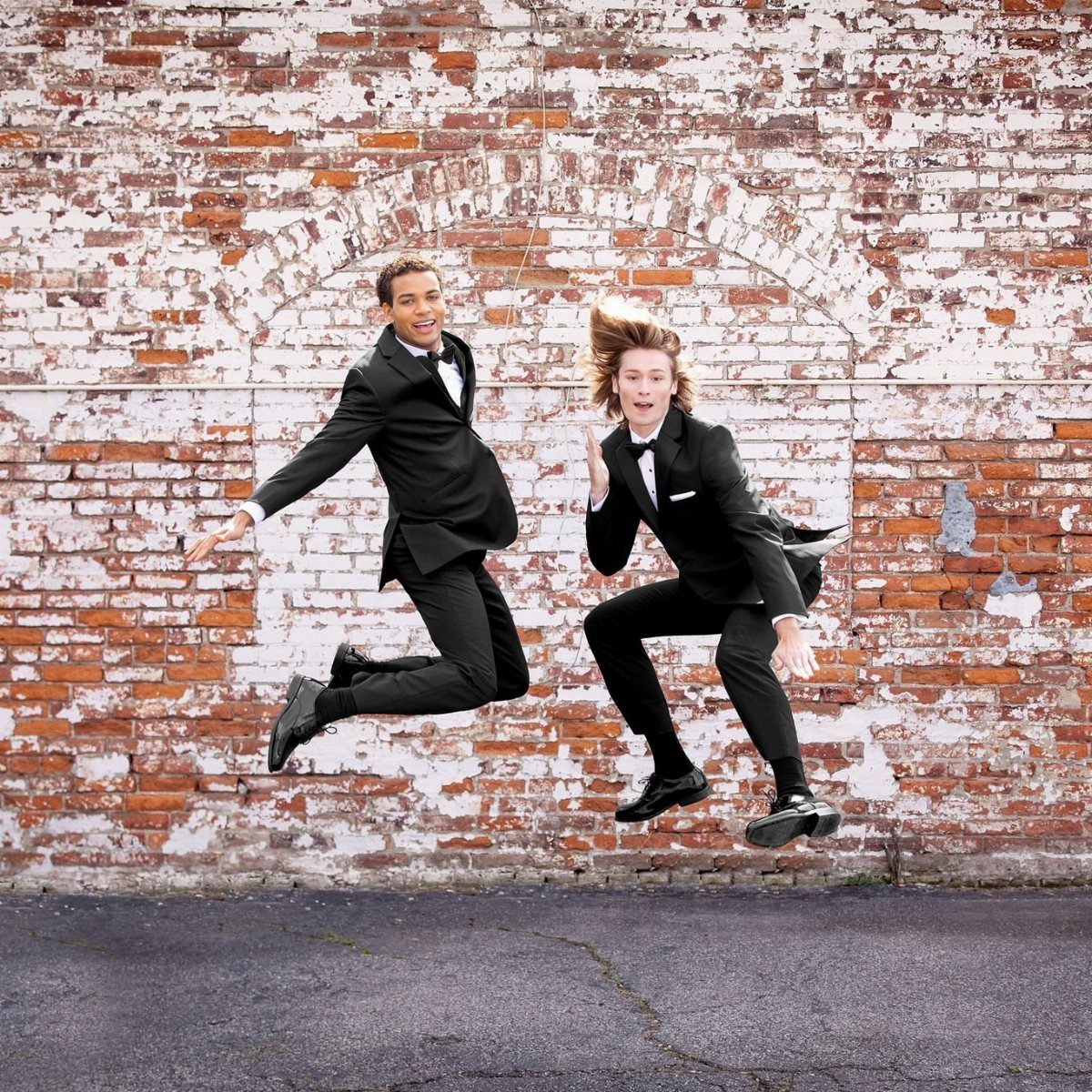 Two young men in black tuxedos jumping excitedly in front of a brick wall.
