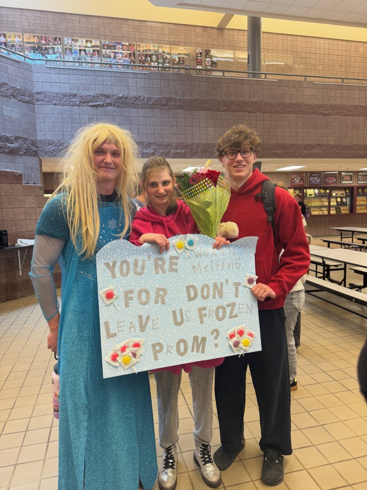 Three teenagers posing with a Frozen-themed prom proposal sign in a school cafeteria.