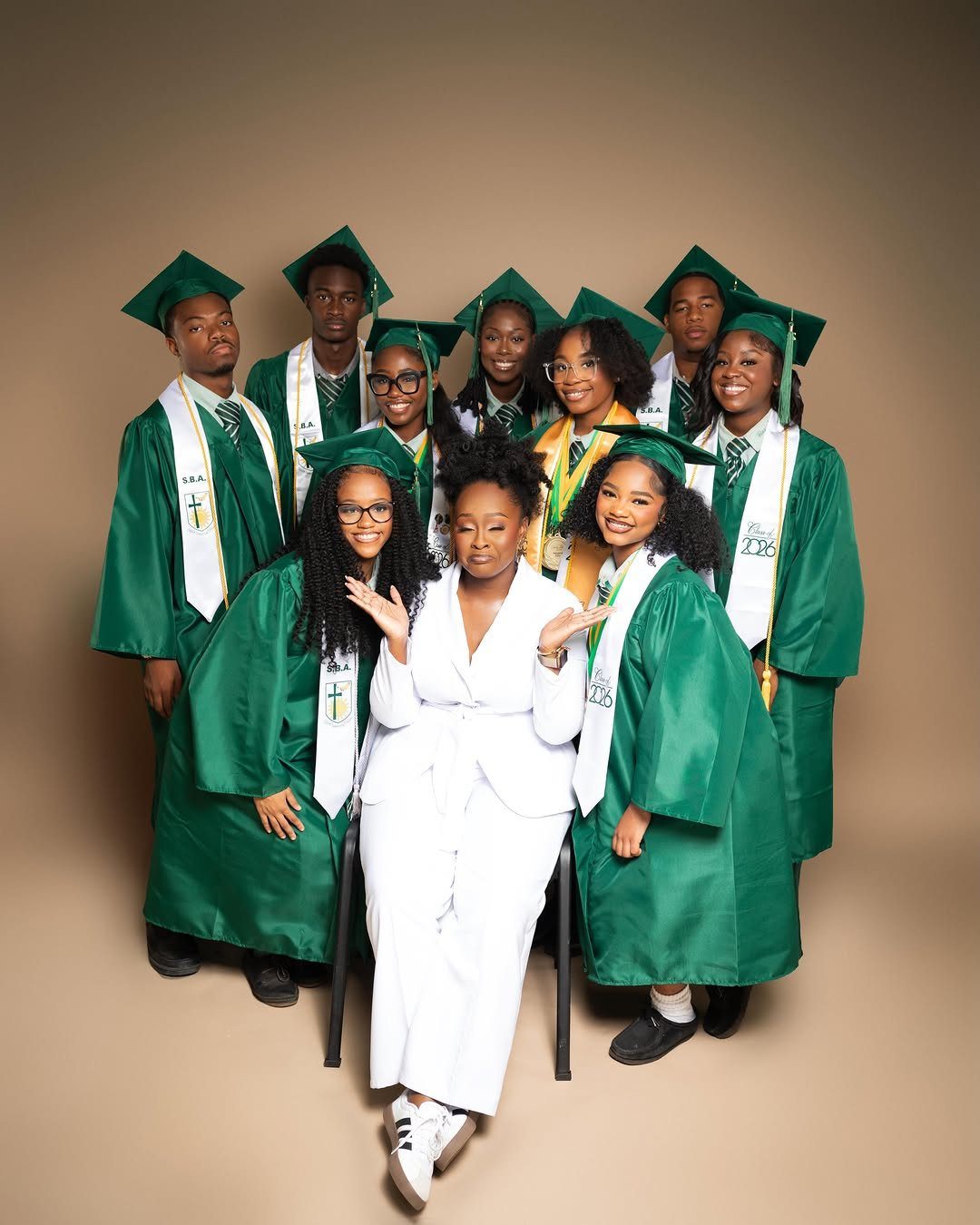 Group of graduates in green gowns posing with a woman in white suit.