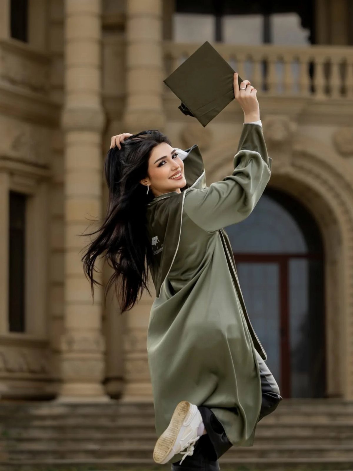 Woman in olive green graduation gown holding a cap before a stone building