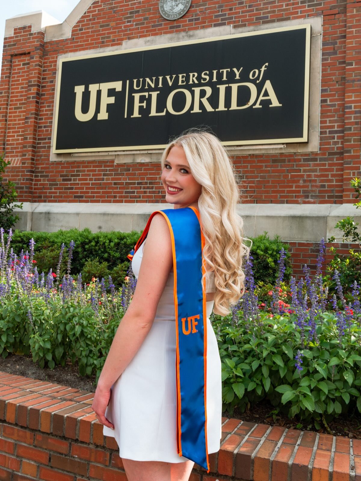 Blonde woman in graduation stole posing before University of Florida sign and flowers.